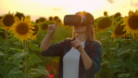 The-girl-farmer-manager-with-long-hair-in-plaid-shirt-and-jeans-is-working-in-VR-glasses.-She-is-engaged-in-the-working-process.-It-is-a-wonderful-summer-evening-in-the-sunflower-field-at-sunset.